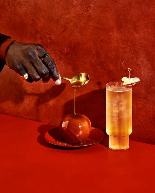 A golden spoon pours liquid over a caramel apple on a red plate, with a cocktail and a hand visible against a textured red background.