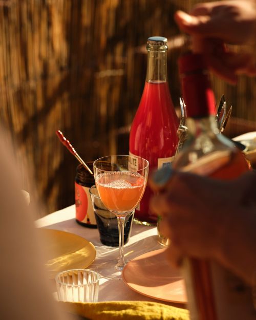 Still life shot of drinks and plates on a table with sunlight highlighting the setting, bottle of pink lemonade, jam, and glassware.
