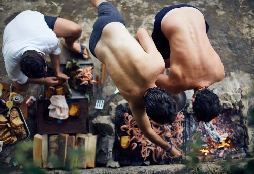 Three men cooking seafood over an outdoor fire pit in a rustic setting. Overhead view captures the grilling and food preparation.