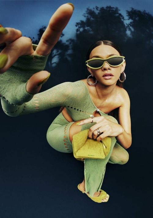 Woman in green patterned outfit and sunglasses crouching forward with hand extended toward the camera against a dark sky background.