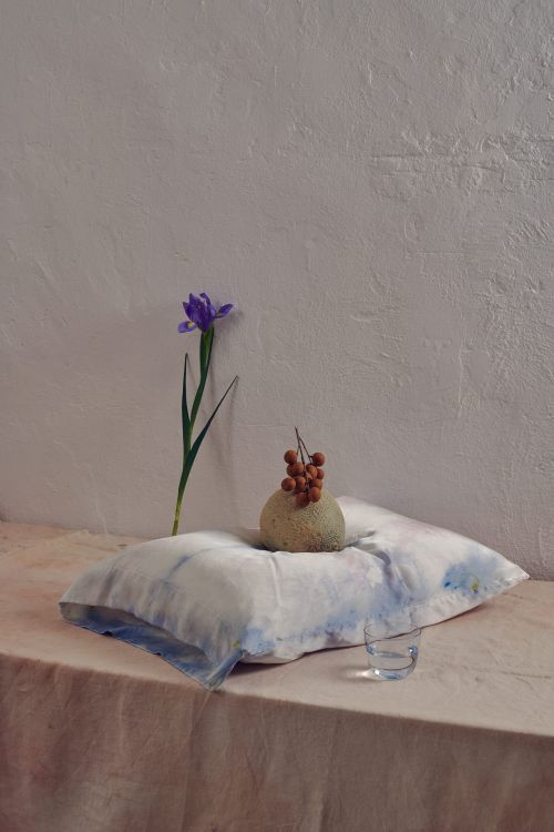 Still life composition with a light-blue pillow, a vase-like fruit, a single violet flower, and a transparent glass in a neutral setting.