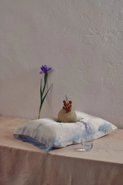 Still life composition with a light-blue pillow, a vase-like fruit, a single violet flower, and a transparent glass in a neutral setting.