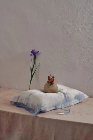 Still life composition with a light-blue pillow, a vase-like fruit, a single violet flower, and a transparent glass in a neutral setting.