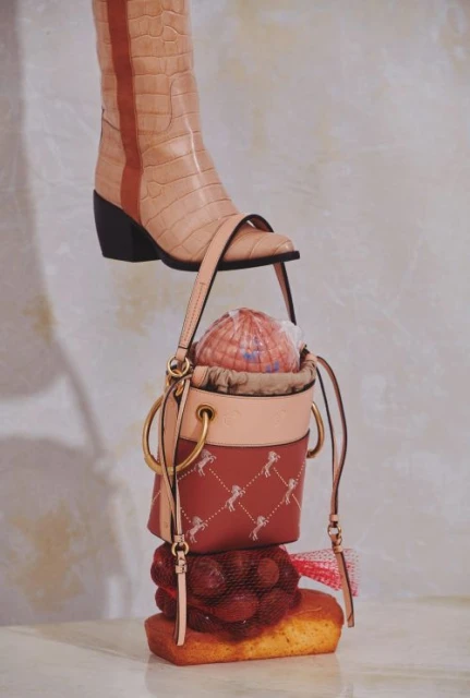 Fashion still life: A beige boot stands atop a bucket bag, resting on cherries and a cake, set against a textured neutral backdrop.