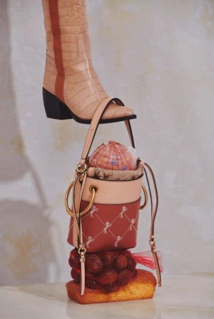 Fashion still life: A beige boot stands atop a bucket bag, resting on cherries and a cake, set against a textured neutral backdrop.