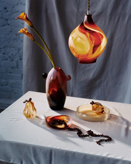 Studio shot featuring glass vase with calla lilies, pendant lamp, and small glass dishes on a white tablecloth, in warm color tones.