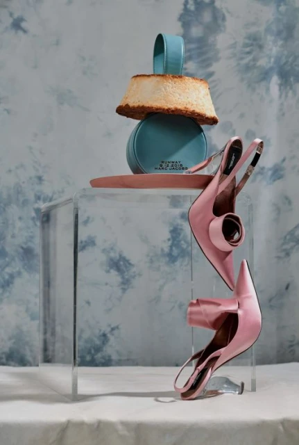 Still life with a pair of pink heels, a cake, and a blue round handbag stacked on a clear acrylic cube against a cloud-patterned backdrop.