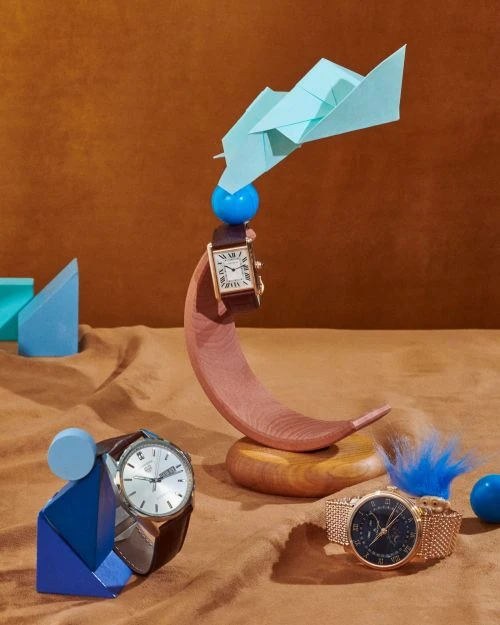 Still life of wristwatches on geometric stands, featuring light blue origami, against a tan backdrop with a shallow depth of field.