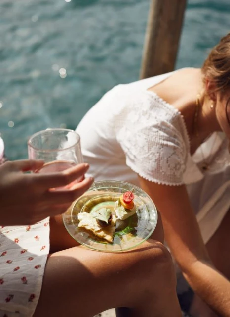 Close-up of a plate of appetizers held by a woman outdoors near water, featuring fresh ingredients, creating an inviting and summery scene.