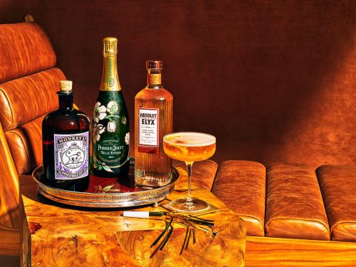 Still life shot of three liquor bottles and a stemmed glass cocktail on a wooden surface, set on a brown leather couch indoors.