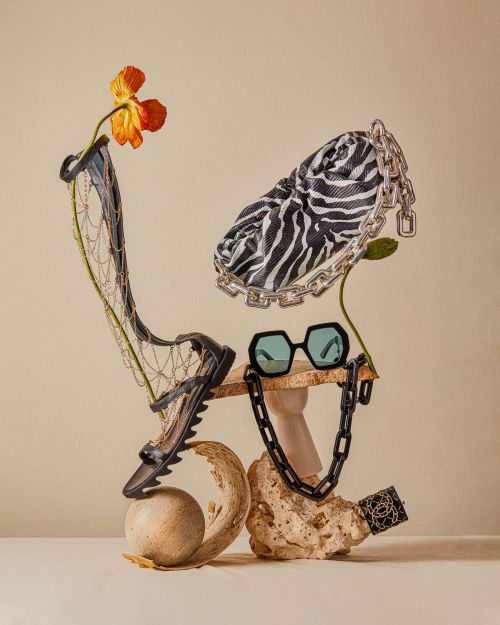 Fashion still life with zebra purse, black sandal, sunglasses, orange flower, and necklace arranged on stone-like platforms.