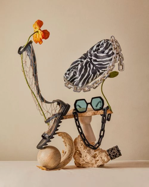 Fashion still life with zebra purse, black sandal, sunglasses, orange flower, and necklace arranged on stone-like platforms.