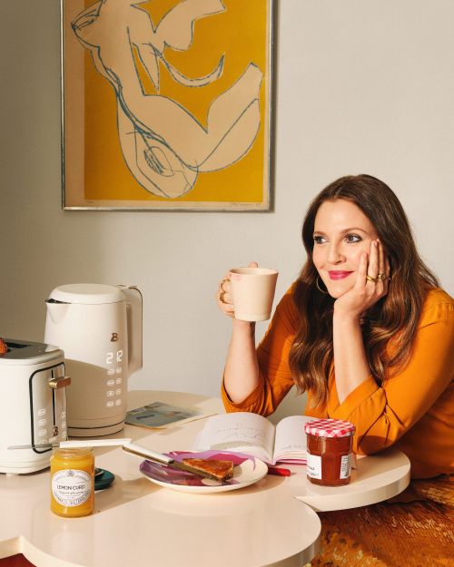 Drew Barrymore sitting at a table with a cup, toaster, kettle, jams, and drawing in frame; yellow, cream, brown, white tones.