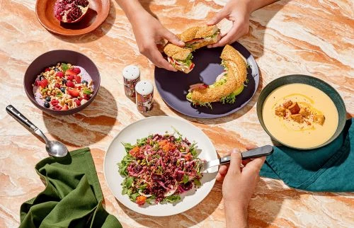 Overhead shot of assorted foods including soup, salad, sandwich, and fruit arranged on a marbled table, with hands reaching in.