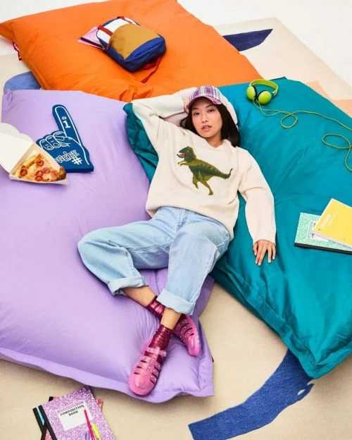 Woman relaxes on colorful pillows with pizza, school supplies, and a backpack in a casual setting, wearing a dinosaur sweater.