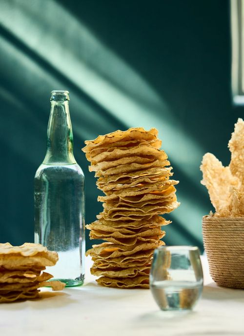 Tall stack of thin round crackers next to a green glass bottle and a woven bowl on a table.