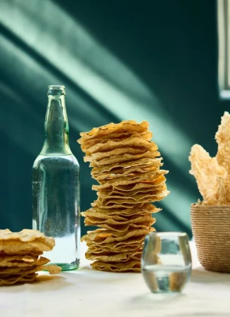 Tall stack of thin round crackers next to a green glass bottle and a woven bowl on a table.