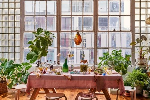 Sunlit dining table with a pink tablecloth, flowers, dishes, and plants in front of a large window in an urban apartment.