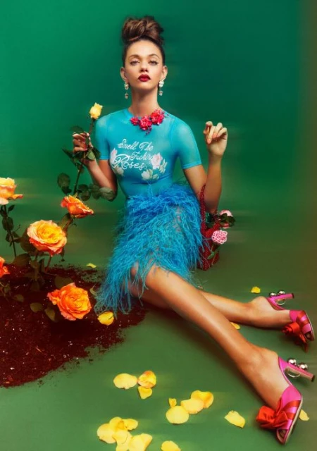 Eye-level studio shot: woman in a blue top and feathered skirt poses near yellow roses, set against a green backdrop with rose petals.