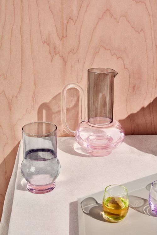Still life of a glass pitcher and various drinking glasses filled with liquids on a white surface against a pink wood background.