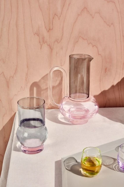 Still life of a glass pitcher and various drinking glasses filled with liquids on a white surface against a pink wood background.