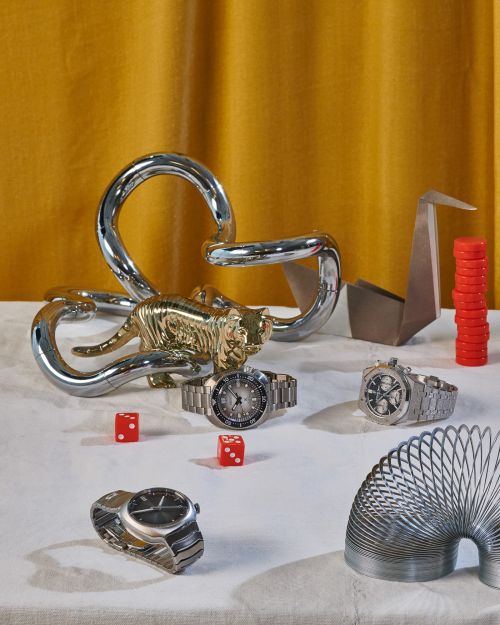 Still life on a white tablecloth features wrist watches, a chrome tiger sculpture, red dice, and other metal and paper objects.