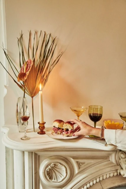 Still life photo of a hand taking a slider from a plate on a white fireplace mantel next to a candle, drinks, and flower arrangement.