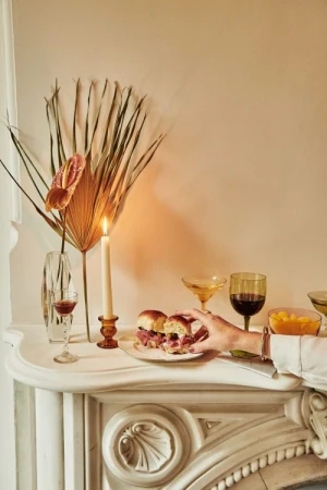 Still life photo of a hand taking a slider from a plate on a white fireplace mantel next to a candle, drinks, and flower arrangement.