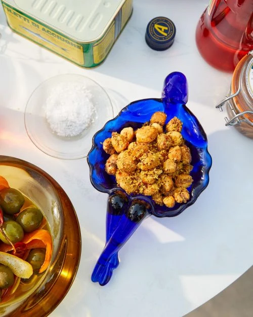 Overhead shot of a blue glass bowl full of nuts, alongside a small bowl of sugar, a tin, olives, and a bottle of liquid.