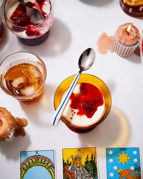 Overhead shot of cocktail, dessert bowls with jam, cupcake candles, tarot cards, spoons on a white surface, studio lighting.
