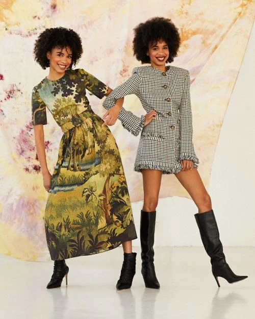 Two smiling adult women model patterned dresses and black boots in a studio with a painterly background. Fashion shoot, full shot.