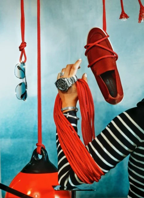 Red casual shoe with silver watch and sunglasses hanging from ropes against a blue-grey background in a studio product shot.