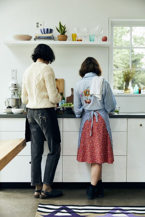 Two women stand at a white kitchen counter preparing food, with one in a floral apron and the other in jeans and a cream-colored sweater.
