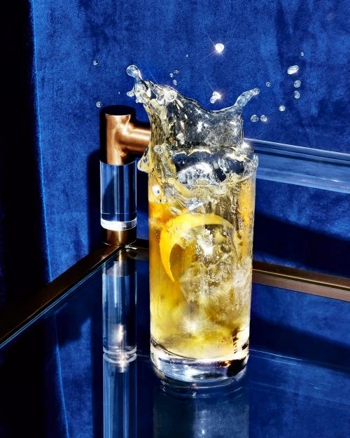 Cocktail splashing in a glass on a glossy blue surface with a blue velvet background and geometric gold metal and acrylic bar accent.