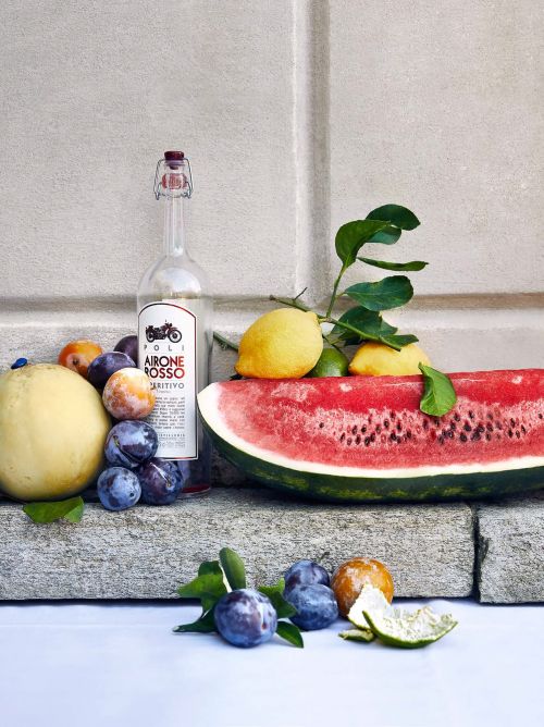 Still life featuring a liquor bottle, watermelon slice, plums, lemon, and melon on a stone surface against a neutral background.
