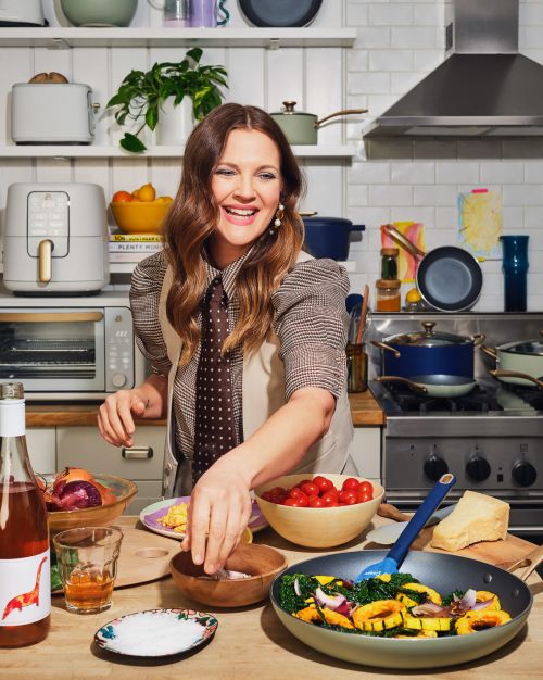 Drew Barrymore in kitchen with various prepared foods, vegetables and pans, wearing a patterned blouse and vest; neutral background.