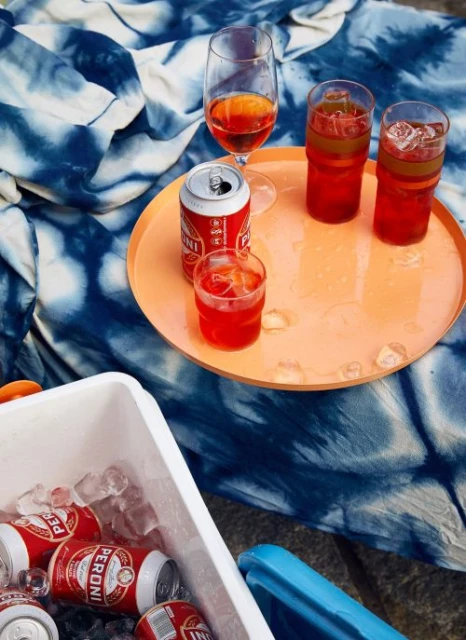 Outdoor picnic with Aperol spritz drinks in glasses and cans, on a blue and white patterned cloth, capturing a relaxed, summery vibe.