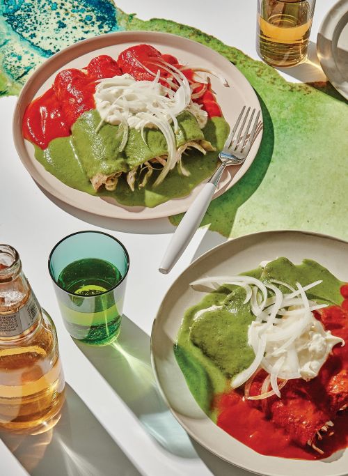 Two plates of enchiladas are arranged on a white surface with a colorful background, accompanied by a green glass and beer.