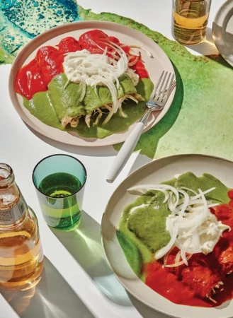 Two plates of enchiladas are arranged on a white surface with a colorful background, accompanied by a green glass and beer.