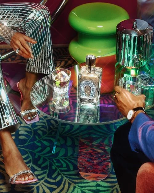 Two people at a table with cocktails, featuring a bottle of tequila, glassware, and shiny disco-inspired clothing in a lounge setting.
