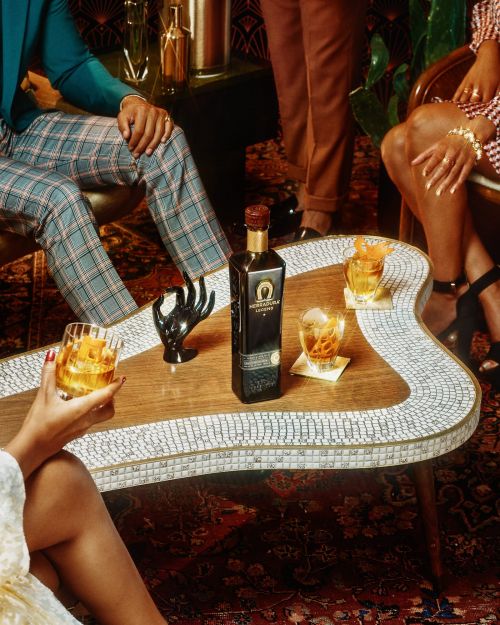 People gather around a coffee table, enjoying drinks with orange peels and featuring a bottle of Peninsula Lounge liquor.