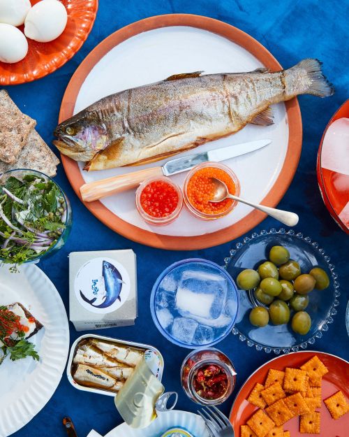 Overhead shot of a cooked fish, caviar, eggs, crackers, and a drink on a blue tablecloth. Food photography showcasing a seafood spread.