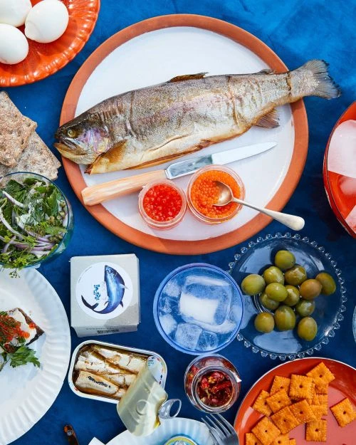 Overhead shot of a cooked fish, caviar, eggs, crackers, and a drink on a blue tablecloth. Food photography showcasing a seafood spread.
