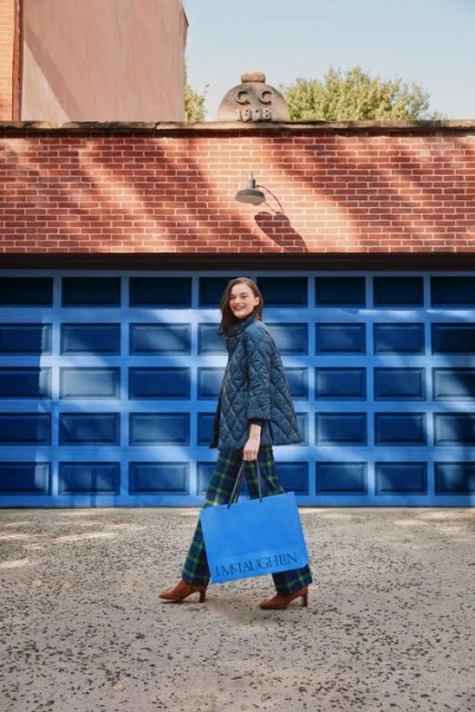 Adult woman in quilted coat and plaid pants walks with a blue shopping bag in front of a blue garage door on an urban street.