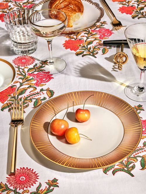 Overhead shot of golden plate with cherries, crystal glasses, pastry, and gold flatware on a white floral patterned tablecloth.