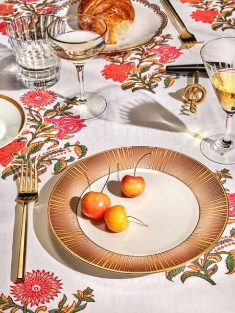 Overhead shot of golden plate with cherries, crystal glasses, pastry, and gold flatware on a white floral patterned tablecloth.