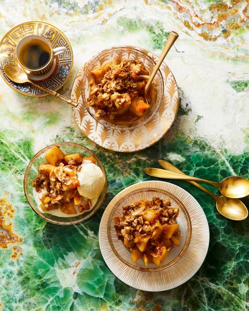 Overhead view of three glass bowls with apple crumble, ice cream, a coffee cup, and gold spoons on a green marble surface.