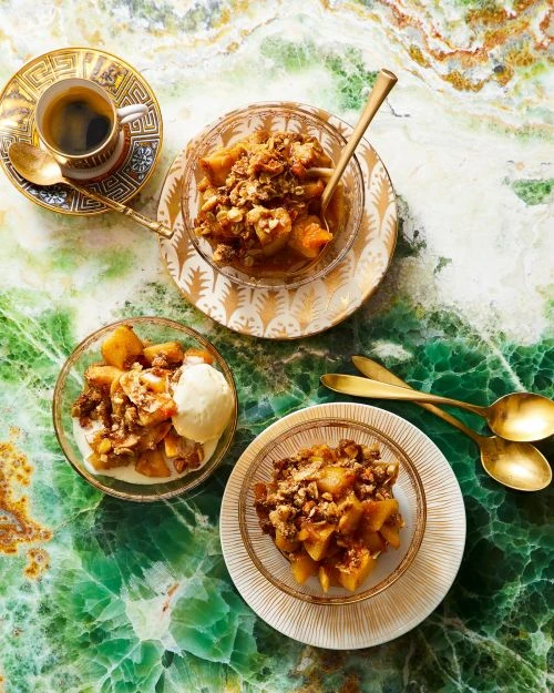 Overhead view of three glass bowls with apple crumble, ice cream, a coffee cup, and gold spoons on a green marble surface.