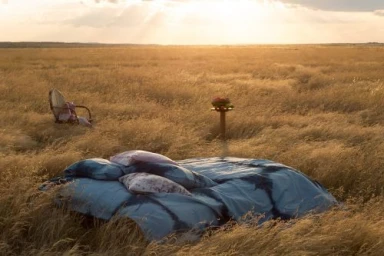 An outdoor bed setup with blue bedding and pillows in the middle of a vast golden meadow under a sunset sky.