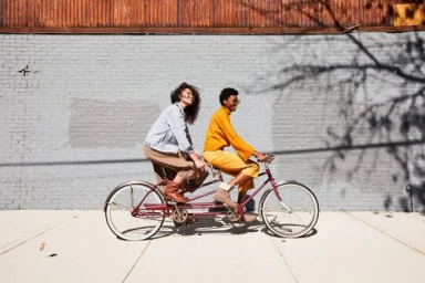 Two smiling women riding a tandem bicycle together along a light grey brick wall in New York, styled by Kalen Kaminski.